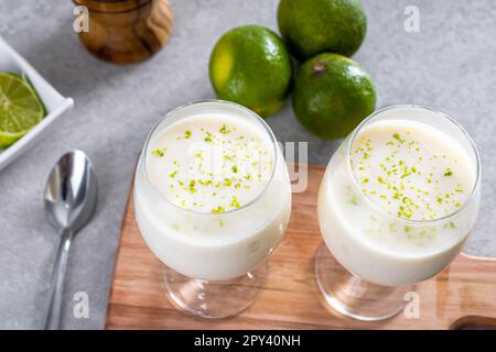 Deliziosa mousse di limone su un asse di legno e uno sfondo bianco da tavolo, con limoni interi e affettati sullo sfondo, e un cucchiaio di metallo. Foto Stock