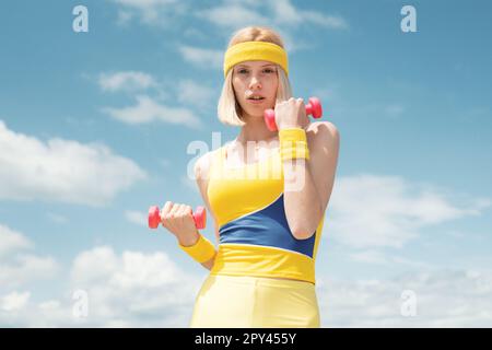 Sano giovane sportivo attraente indossando giallo con uniforme blu di allenamento dimostrazione esercizi con manubri rosa su sfondo cielo con Foto Stock