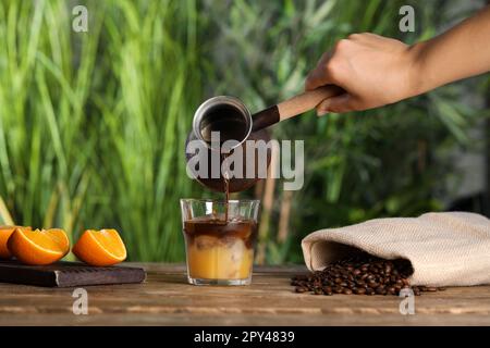 Donna che versa il caffè in vetro con succo d'arancia e cubetti di ghiaccio al tavolo di legno, primo piano Foto Stock