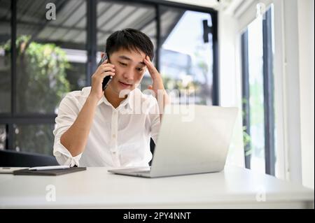 Sottolineato e premuroso uomo d'affari asiatico millenario che si occupa di affari con il suo cliente aziendale al telefono, mentre seduto alla sua scrivania. Foto Stock