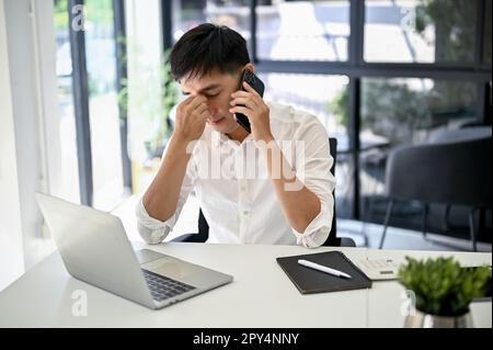 Sottolineato e premuroso uomo d'affari asiatico millenario parlando al telefono con il suo lavoratore e risolvere i problemi al telefono. Foto Stock