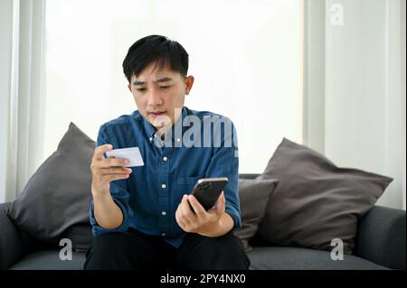 Ha sottolineato l'uomo asiatico millenario che ha un problema con la sua applicazione di mobile banking, tenendo il telefono cellulare e guardando la sua carta di credito con faccia seria Foto Stock