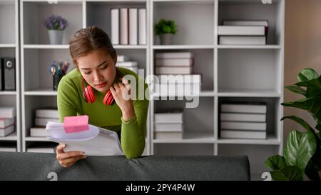 Donna d'affari asiatica millenaria focalizzata in abiti casual appoggiata su un divano nel suo ufficio e leggendo documenti. Foto Stock