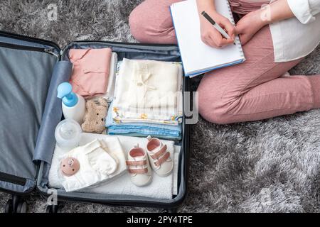 Vista dall'alto della donna incinta che scrive la lista di imballaggio per l'ospedale di maternità seduto accanto alla valigia Foto Stock