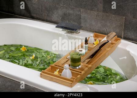 Vasca piena di acqua ed erbe con vassoio da bagno in legno con vari prodotti cosmetici, relax e benessere concetto di stile di vita Foto Stock