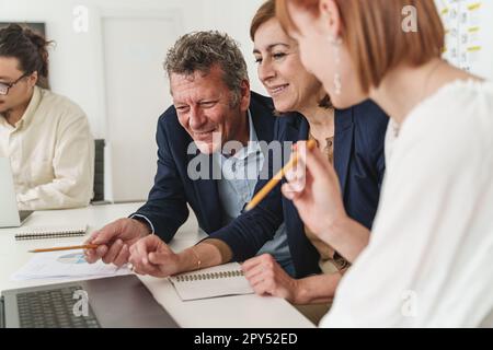 In un ufficio moderno, tre colleghi caucasici vestiti casualmente, due anziani e una giovane donna, si concentrano sullo schermo di un notebook, mentre un giovane asiatico lavora Foto Stock