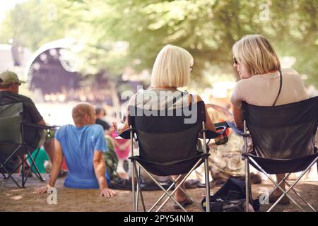 Ritorno, amici seduti e festival all'aperto su sedia da campeggio, parlare e le donne in pubblico, concerto o spettacolo musicale. Persone, tifosi rilassante Foto Stock