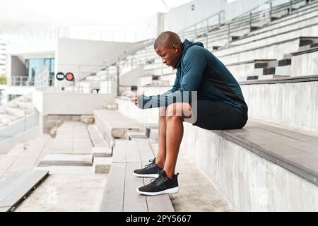 Risposta ai miei messaggi di testo. Scatto a tutta lunghezza di un bel giovane atleta seduto da solo e SMS sul suo cellulare dopo un allenamento all'aperto Foto Stock