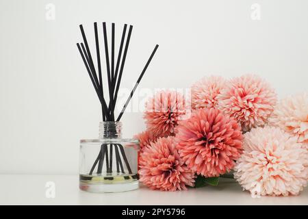 Composizione con bastoncini di incenso, olio essenziale in vaso di vetro e fiori artificiali di spugna rosa all'interno di una stanza bianca. Sfondo bianco. Molti fiori e bastoni profumati. Foto Stock