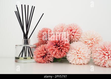 Composizione con bastoncini di incenso, olio essenziale in vaso di vetro e fiori artificiali di spugna rosa all'interno di una stanza bianca. Sfondo bianco. Molti fiori e bastoni profumati. Foto Stock