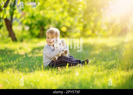 Un ragazzino carino seduto sul prato in giardino e un soffice giocattolo. Foto Stock