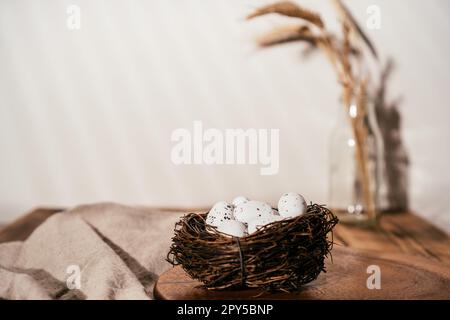 Composizione pasquale di uova di quaglia in nido su tavola di legno e rametto di frumento in vetro su sfondo bianco beige. Foto Stock