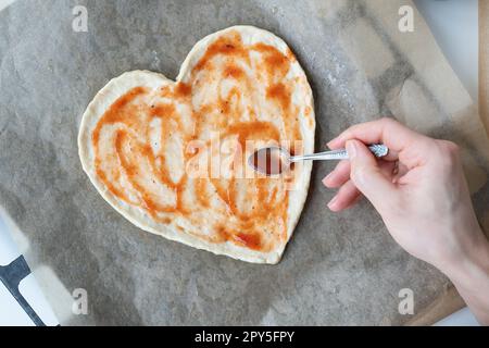 Impasto della pizza a forma di cuore, lo chef spalma la salsa di pomodoro sulla pizza. Il concetto di sorpresa per St. San Valentino. Foto Stock