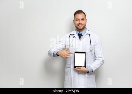 Giovane medico caucasico con capelli corti, baffi e barba vestita di abito bianco e stetoscopio che punta il dito verso il monitor del computer tablet. Posizionatevi di fronte alla parete bianca. Foto Stock