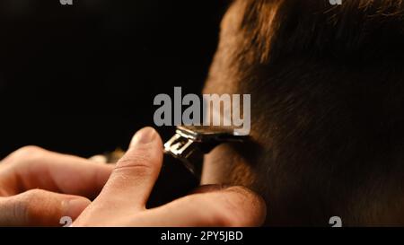 Immagine ravvicinata di un bel barbiere che taglia i capelli al suo cliente usando il trimmer. Servizio di parrucchiere in un moderno barbiere con illuminazione a chiave scura e calda vista sul retro Foto Stock