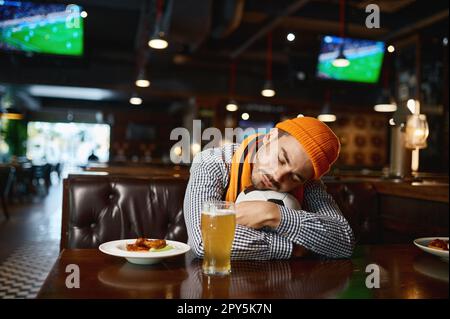 Divertente giovane uomo tifoso di calcio che dorme sulla palla mentre riposa nello sport bar Foto Stock