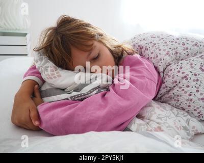 Una bella ragazza caucasica di 8 anni con capelli biondi, vestita con pigiama rosa, dorme su un letto con una soffice coperta, abbracciando un cuscino. La luce soffusa del sole del mattino scorre attraverso la finestra. Foto Stock