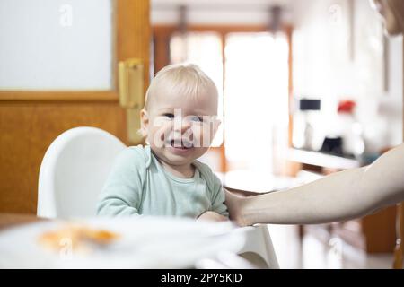 Adorabile allegro bambino ragazzo felice bambino sorridente mentre si siede in seggiolone al tavolo da pranzo in cucina a casa beeing cucchiaio alimentato da sua madre. Foto Stock