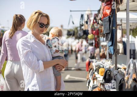 La madre che tiene il suo bambino bambino bambino piccolino che frusta oggetti al mercatino delle pulci della domenica. Foto Stock