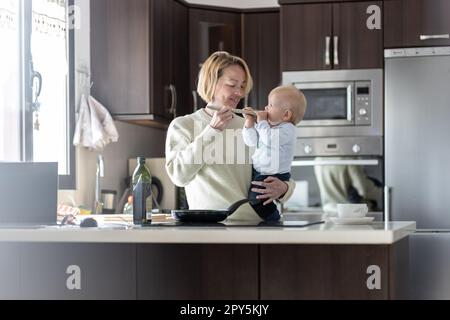 Mamma felice e bambino piccolo cucino cucinare e assaggiare una cena sana in cucina domestica. Famiglia, stile di vita, vita domestica, cibo, alimentazione sana e concetto di persone. Foto Stock