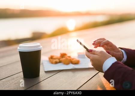 Primo piano delle mani femminili che usano lo smartphone la sera sul fiume. Donna che scrive messaggi sul social network nel parco con il tramonto sullo sfondo, caffè da asporto e pepite da fast food sullo sfondo Foto Stock