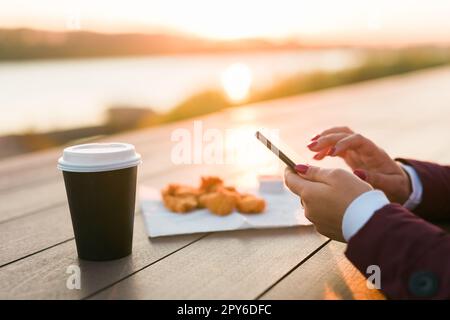 Primo piano delle mani femminili che usano lo smartphone la sera sul fiume. Donna che scrive messaggi sul social network nel parco con il tramonto sullo sfondo, caffè da asporto e pepite da fast food sullo sfondo Foto Stock