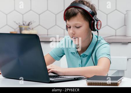 Ritratto di un giovane preadolescente che indossa cuffie, seduto alla scrivania vicino a notebook, che usa un computer portatile, che tiene lezioni online. Foto Stock