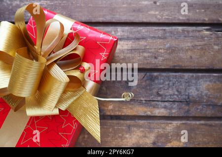 Confezione regalo rossa con grande fiocco dorato su sfondo in legno. Regalo per Natale, Capodanno, compleanno, festa della mamma, matrimonio, San Valentino. Bella decorazione festiva. Prua lussureggiante fatta di nastro giallo. Foto Stock