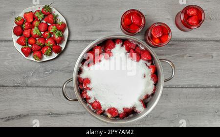 Il piano portapaziente foto - grande pentola in acciaio di fragole ricoperte con cristalli di zucchero, piastra e bottiglie in vetro con più frutti attorno al grigio scrivania di legno. Foto Stock