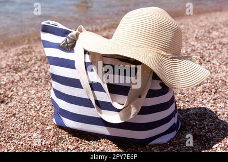 Borsa accessori spiaggia e cappello sulla spiaggia di ciottoli vicino al mare. Sfondo estivo. Foto Stock