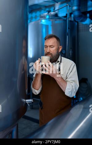 Birraio assaggia birra fresca mentre si trova davanti ai contenitori di metallo Foto Stock