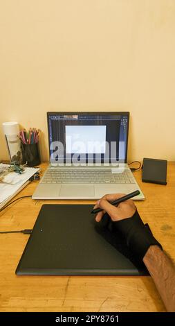 un designer che lavora sul suo notebook utilizzando un tablet per schizzi e una penna digitale per il suo lavoro di grafica creativa sul tavolo di lavoro con la penna a colori Foto Stock