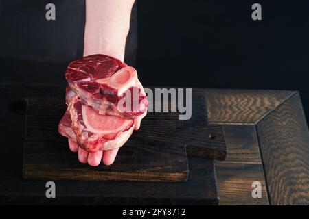 Lo chef taglia una pila di carne di manzo da bistecche diverse o diverse. Le mani di Mans tengono la bistecca cruda della pila sul tagliere di legno rustico su sfondo nero. Foto Stock