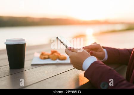 Primo piano delle mani femminili che usano lo smartphone la sera sul fiume. Donna che scrive messaggi sul social network nel parco con il tramonto sullo sfondo, caffè da asporto e pepite da fast food sullo sfondo Foto Stock