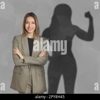 Donna d'affari e ombra di forte signora muscolare dietro di lei sulla parete grigia. Concetto di resistenza interna Foto Stock