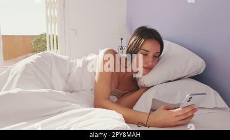 Donna felice che SMS al telefono dopo il risveglio al mattino. Ragazza sorridente che ha una conversazione online, rispondendo al messaggio TAP mentre si siede a letto Foto Stock