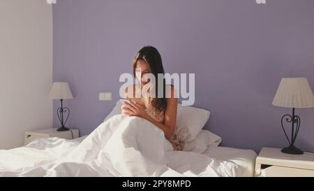 Donna felice che SMS al telefono dopo il risveglio al mattino. Ragazza sorridente che ha una conversazione online, rispondendo al messaggio TAP mentre si siede a letto Foto Stock