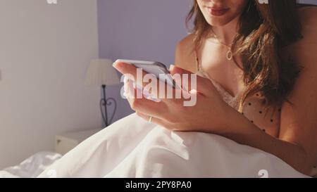 Donna felice che SMS al telefono dopo il risveglio al mattino. Ragazza sorridente che ha una conversazione online, rispondendo al messaggio TAP mentre si siede a letto Foto Stock