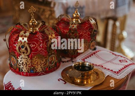 Vista in primo piano di due corone rosse di nozze su asciugamano ricamato e ciotola con acqua santificata durante una cerimonia nuziale nella chiesa ortodossa Ucraina. Foto Stock