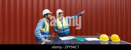 Addetto alla spedizione che indica la posizione di stoccaggio del contenitore, spiegare al collega la pianificazione della spedizione successiva. Usano il pannello delle celle solari come tavolo. Sullo sfondo è presente un contenitore di grandi dimensioni Foto Stock