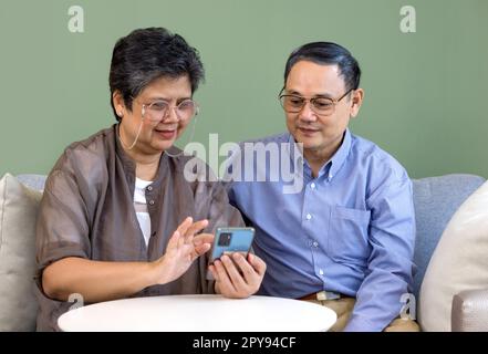 Le coppie asiatiche trascorrono del tempo insieme in soggiorno. Donna e uomo in pensione che guardano una foto scattata dal telefono cellulare. Stile di vita moderno relazioni e attività tra le persone. Foto Stock