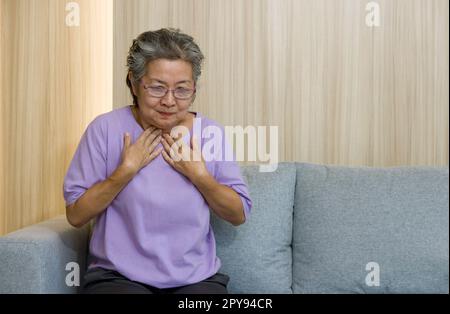 Anziani asiatici con gli occhiali, posare entrambe le mani sul petto a causa della nausea mentre si siede sul divano nel soggiorno. Foto Stock