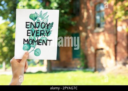 Donna che tiene la carta con frase godere ogni momento all'aperto, primo piano. Spazio per il testo Foto Stock