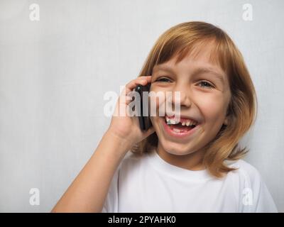 bambina di 7 anni senza dente anteriore. Il bambino sta parlando al telefono o allo smartphone e ride allegramente. Cambio dei denti del latte nell'infanzia. Moderne tecnologie di comunicazione. Panno bianco su una ragazza Foto Stock