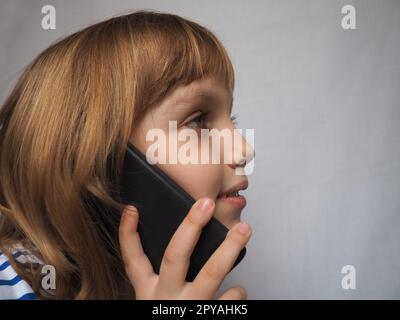 bambina con i capelli biondi che parla su smartphone, telefono o tablet. Sviluppo di tecnologie e telecomunicazioni, loro impatto sulla salute. Stile di vita moderno. Bel bambino di 7 anni Foto Stock