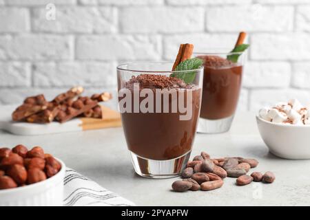 Tazze di delizioso cioccolato caldo con bastoncino di cannella, menta e fagioli di cacao su un tavolo grigio chiaro vicino al muro di mattoni Foto Stock