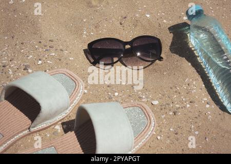 Eleganti occhiali da sole, ciabatte e bottiglia d'acqua sulla sabbia Foto Stock