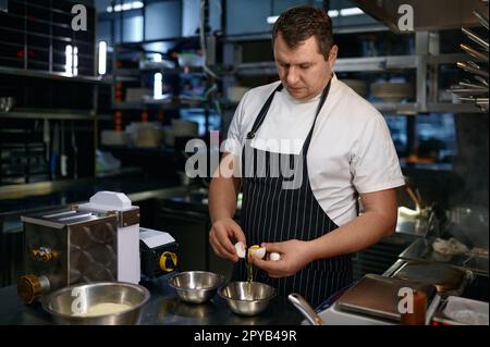 Chef maturo in grembiule cracking uova fresche, mentre cucinare noodle Foto Stock