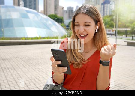 Una giovane donna entusiasta che urla ascoltando la sua canzone preferita su un'applicazione musicale dello smartphone utilizzando gli auricolari wireless in una metropoli moderna di mattina Foto Stock