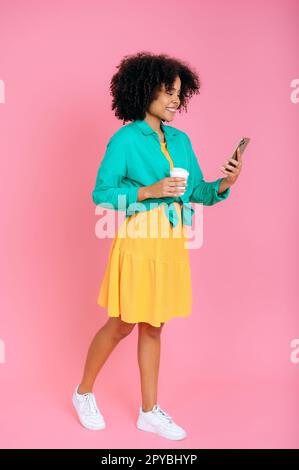 Foto a tutta lunghezza di una bella ragazza con capelli ricci e abiti eleganti utilizzando il suo smartphone per la messaggistica sui social media, bere caffè, stare in piedi su sfondo rosa isolato, sorridere Foto Stock
